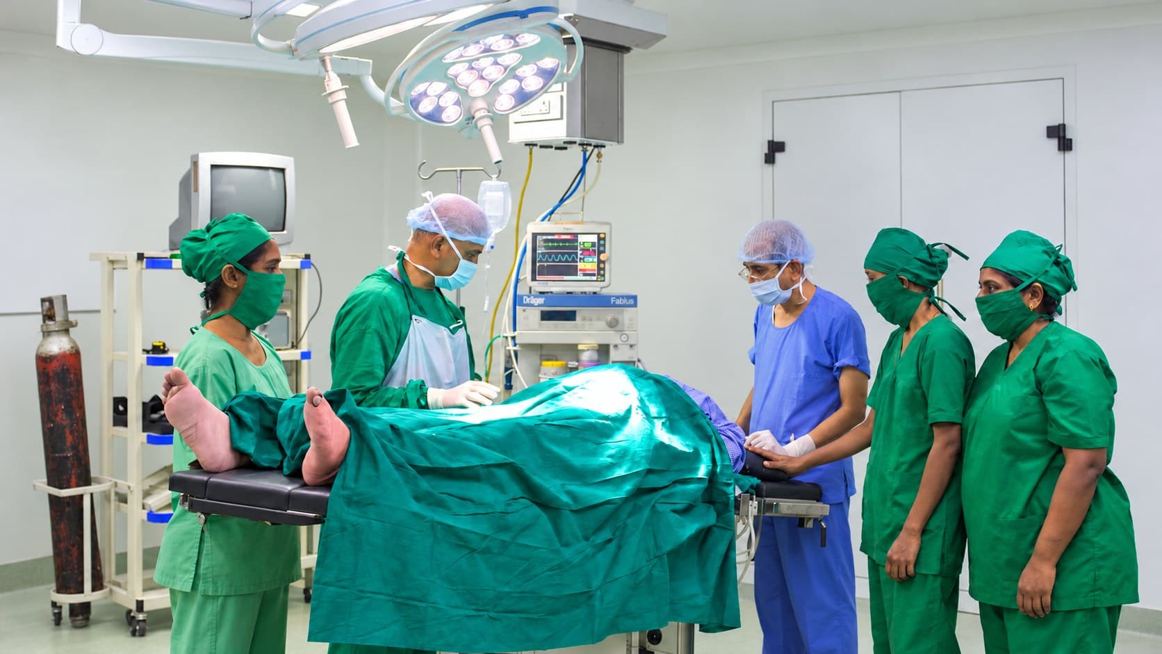 A surgical team in green and blue scrubs at the operating table under an LED theatre light, with a Dräger Fabius anaesthesia workstation behind them