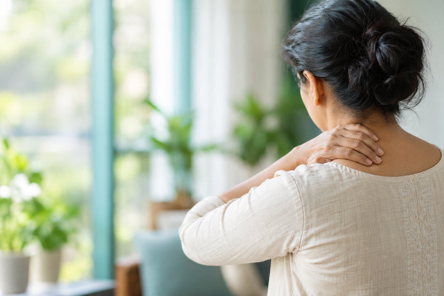 A woman holding the back of her stiff neck, seen from behind in a bright room with plants